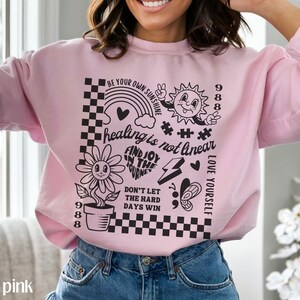 May include: Light pink sweatshirt with a black graphic print featuring a flower, sun, rainbow, and the text "healing is not linear", "FIND JOY IN THE JOURNEY", "DON'T LET THE HARD DAYS WIN", "LOVE YOURSELF", and "BE YOUR OWN SUNSHINE".