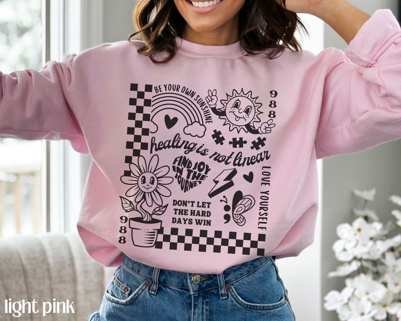 May include: Light pink sweatshirt with a black graphic print featuring a flower, sun, rainbow, and the text "healing is not linear", "FIND JOY IN THE JOURNEY", "DON'T LET THE HARD DAYS WIN", "LOVE YOURSELF", and "BE YOUR OWN SUNSHINE".
