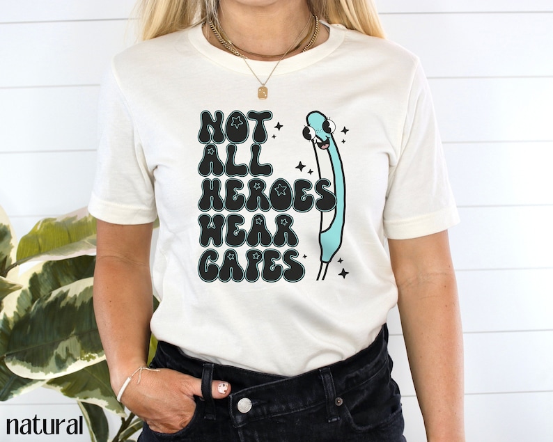 May include: A white t-shirt with a graphic print that says "Not All Heroes Wear Capes" in black with a blue and white cartoon character. The character is a stick figure with a large head and a blue body. The character is wearing a cape and has a star on its head.