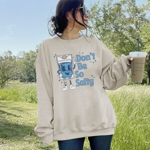 Op de afbeelding: Een beige sweatshirt met een blauw cartoonfiguur dat een zak met zoutoplossing vasthoudt. Het figuur draagt een blauwe halo en de tekst "Don't Be So Salty" is op het sweatshirt gedrukt.