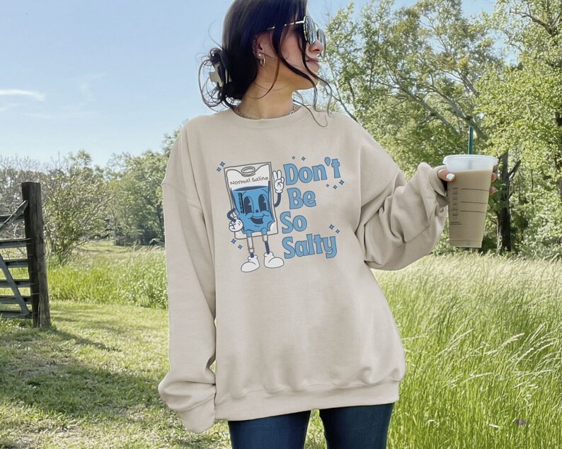 Op de afbeelding: Een beige sweatshirt met een blauw cartoonfiguur dat een zak met zoutoplossing vasthoudt. Het figuur draagt een blauwe halo en de tekst "Don't Be So Salty" is op het sweatshirt gedrukt.