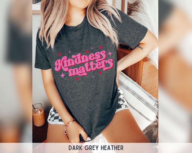 Pode incluir: T-shirt cinza escuro mesclado com o texto "Kindness matters" em rosa e vermelho. O texto tem uma fonte retr&ocirc; e &eacute; rodeado por estrelas. A camisa &eacute; de manga curta.