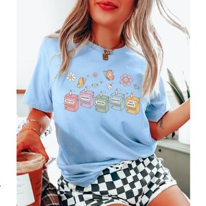 May include: Light blue t-shirt with a graphic of colorful juice boxes with positive affirmations. The text on the boxes reads "Self Love", "Good Vibes", "Empathy", "Inspiration", and "Anti Anxiety".
