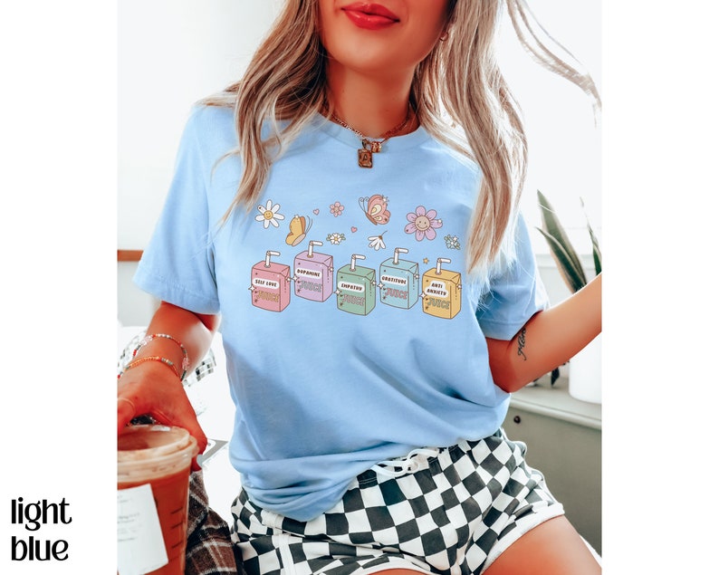May include: Light blue t-shirt with a graphic of colorful juice boxes with positive affirmations. The text on the boxes reads "Self Love", "Good Vibes", "Empathy", "Inspiration", and "Anti Anxiety".