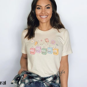May include: A light beige t-shirt with a graphic of six colorful juice boxes with the words "Stay True", "Sunshine", "Kindness", "Gratitude", "Good Vibes", and "Self Love" printed on them. The graphic is surrounded by small flowers and butterflies.