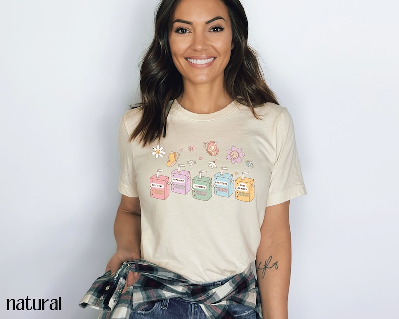 May include: A light beige t-shirt with a graphic of six colorful juice boxes with the words "Stay True", "Sunshine", "Kindness", "Gratitude", "Good Vibes", and "Self Love" printed on them. The graphic is surrounded by small flowers and butterflies.
