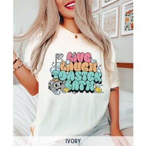 Pu&ograve; includere: Una maglietta bianca con una grafica colorata che dice "Live Laugh Take a Bath". La grafica presenta una doccia, bolle e un'anatra di gomma.