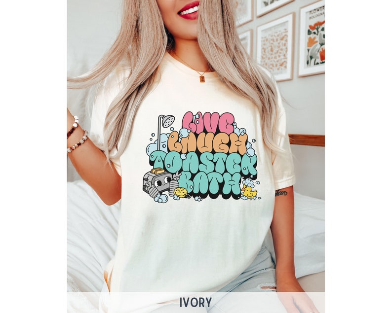 Pu&ograve; includere: Una maglietta bianca con una grafica colorata che dice "Live Laugh Take a Bath". La grafica presenta una doccia, bolle e un'anatra di gomma.