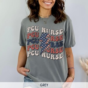 K&ouml;nnte beinhalten: Ein graues T-Shirt mit einem Grafikdesign eines Blitzes mit roten, wei&szlig;en und blauen Streifen. Der Text "PCU NURSE" wird in einem gestapelten Muster um den Blitz herum wiederholt.