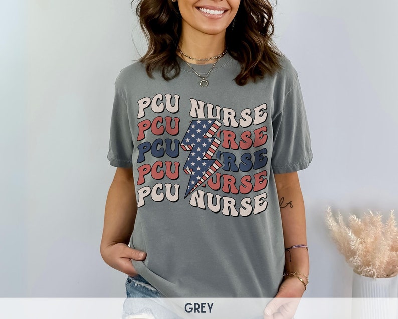 K&ouml;nnte beinhalten: Ein graues T-Shirt mit einem Grafikdesign eines Blitzes mit roten, wei&szlig;en und blauen Streifen. Der Text "PCU NURSE" wird in einem gestapelten Muster um den Blitz herum wiederholt.