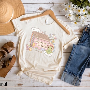 May include: A beige t-shirt with a graphic of a frog holding a puzzle piece and a cassette tape with the text "Close some tabs, take a breath". The graphic is surrounded by flowers and clouds.