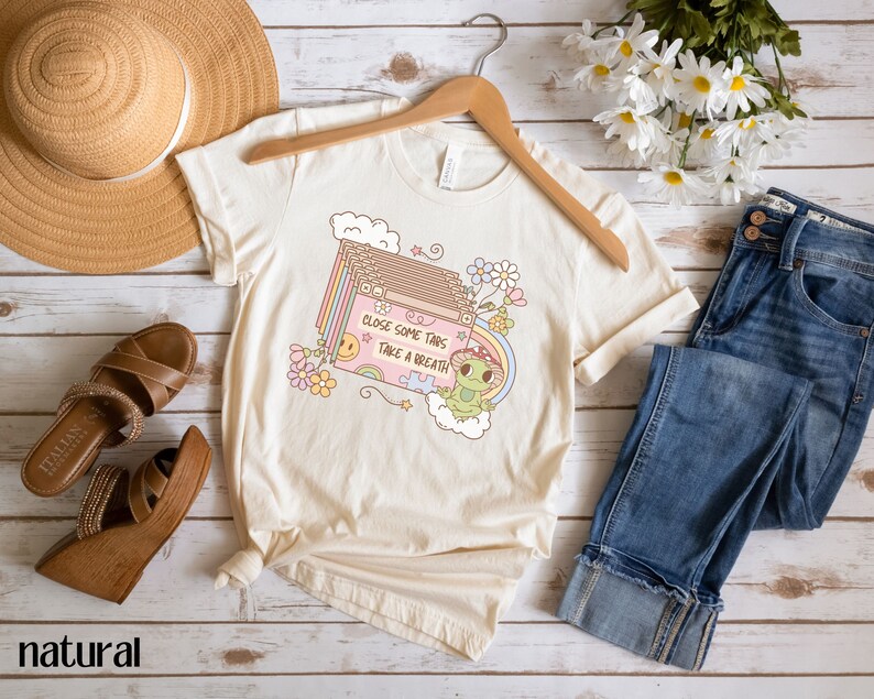 May include: A beige t-shirt with a graphic of a frog holding a puzzle piece and a cassette tape with the text "Close some tabs, take a breath". The graphic is surrounded by flowers and clouds.