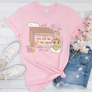 May include: Pink t-shirt with a graphic of a frog wearing a mushroom hat and a stack of browser tabs with the text "Close some tabs, take a breath".