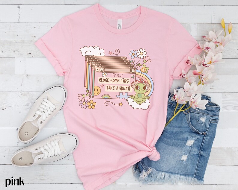 May include: Pink t-shirt with a graphic of a frog wearing a mushroom hat and a stack of browser tabs with the text "Close some tabs, take a breath".