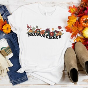 May include: White t-shirt with a floral design and the word "NEUROSCIENCE" in black lettering. The shirt is laid out on a wooden surface with jeans, a coffee cup, and fall foliage.