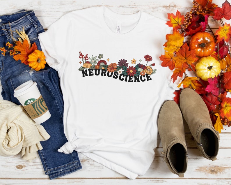 May include: White t-shirt with a floral design and the word "NEUROSCIENCE" in black lettering. The shirt is laid out on a wooden surface with jeans, a coffee cup, and fall foliage.