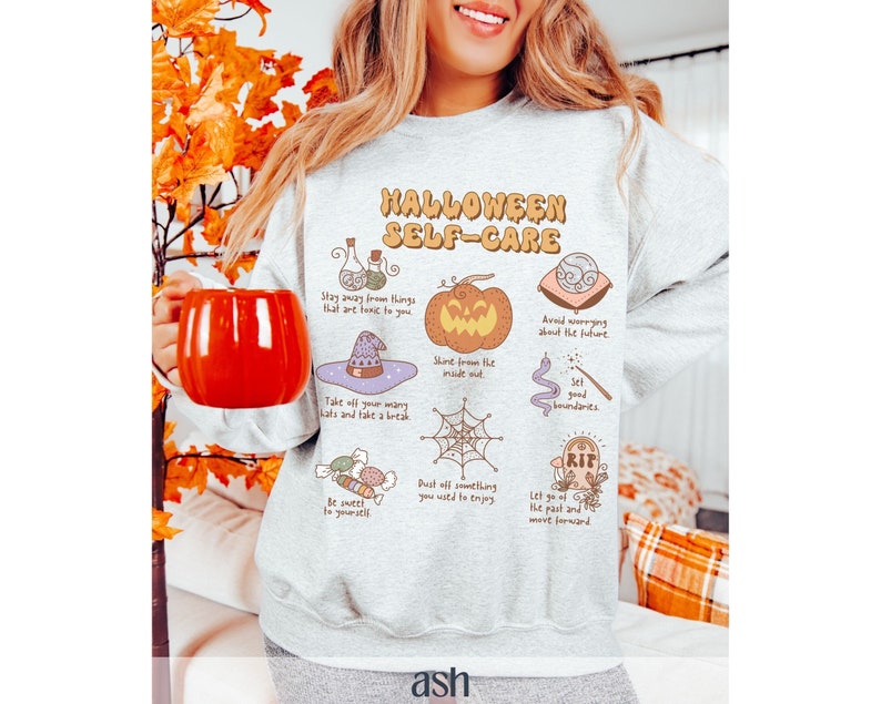 May include: A gray sweatshirt with a Halloween-themed design featuring illustrations of a pumpkin, a witch's hat, a spiderweb, and a tombstone. The text "Halloween Self-Care" is printed in orange and black letters.