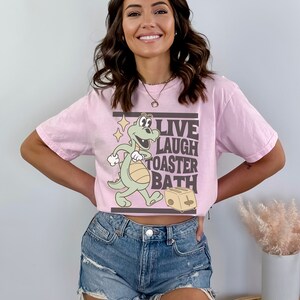 May include: A light pink t-shirt featuring a cartoon dinosaur with the words "LIVE LAUGH TOASTER BATH" in a rectangular frame. The dinosaur is light green with a white belly and is walking towards a toaster.