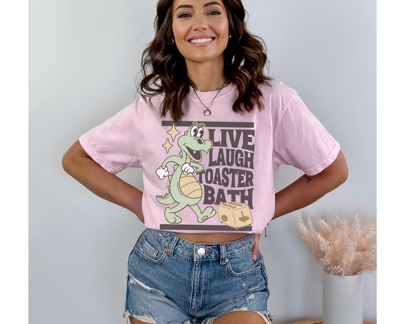 May include: A light pink t-shirt featuring a cartoon dinosaur with the words "LIVE LAUGH TOASTER BATH" in a rectangular frame. The dinosaur is light green with a white belly and is walking towards a toaster.