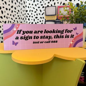 Puede incluir: Un letrero rectangular con el texto "If you are looking for a sign to stay, this is it text or call 988" en blanco. El letrero tiene un fondo rosa con un patr&oacute;n de cuadr&iacute;cula, mariposas y un dise&ntilde;o de arco&iacute;ris.