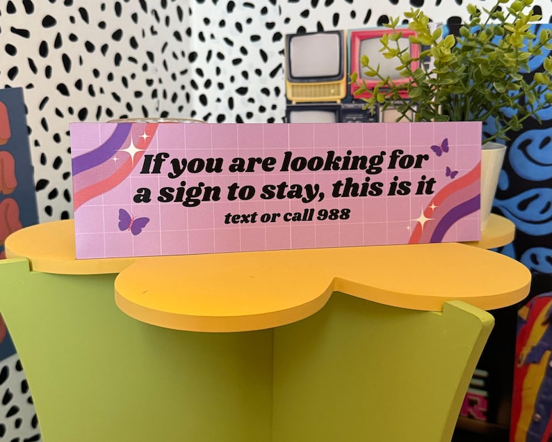 Puede incluir: Un letrero rectangular con el texto "If you are looking for a sign to stay, this is it text or call 988" en blanco. El letrero tiene un fondo rosa con un patr&oacute;n de cuadr&iacute;cula, mariposas y un dise&ntilde;o de arco&iacute;ris.