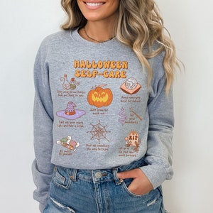 May include: Gray sweatshirt with a Halloween self-care graphic. The graphic features a pumpkin, a witch's hat, a spiderweb, candy, a potion bottle, a tombstone, and a crystal ball. The text reads "Halloween Self-Care".