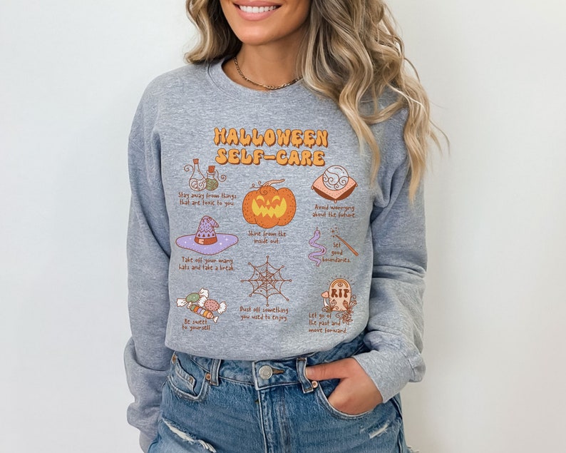 May include: Gray sweatshirt with a Halloween self-care graphic. The graphic features a pumpkin, a witch's hat, a spiderweb, candy, a potion bottle, a tombstone, and a crystal ball. The text reads "Halloween Self-Care".