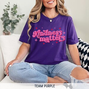 Pode incluir: Uma camiseta roxa com o texto "Kindness matters" em uma fonte retr&ocirc;. O texto &eacute; em rosa e vermelho com detalhes de estrela. A camisa &eacute; usada com shorts jeans azuis.