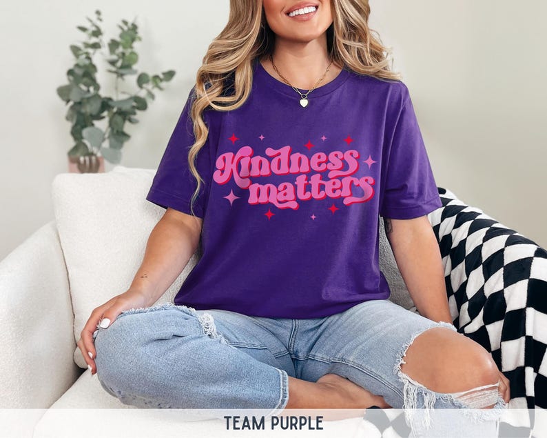 Pode incluir: Uma camiseta roxa com o texto "Kindness matters" em uma fonte retr&ocirc;. O texto &eacute; em rosa e vermelho com detalhes de estrela. A camisa &eacute; usada com shorts jeans azuis.