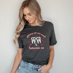 Puede incluir: Una camiseta gris oscuro con un gr&aacute;fico de dos dientes de dibujos animados tomados de la mano con un coraz&oacute;n encima. El texto "Nothing will ever come between us" est&aacute; impreso debajo de los dientes.