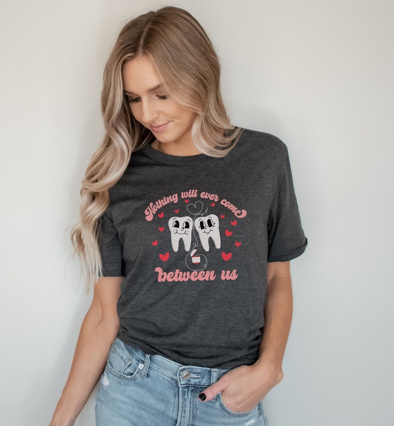 Puede incluir: Una camiseta gris oscuro con un gr&aacute;fico de dos dientes de dibujos animados tomados de la mano con un coraz&oacute;n encima. El texto "Nothing will ever come between us" est&aacute; impreso debajo de los dientes.