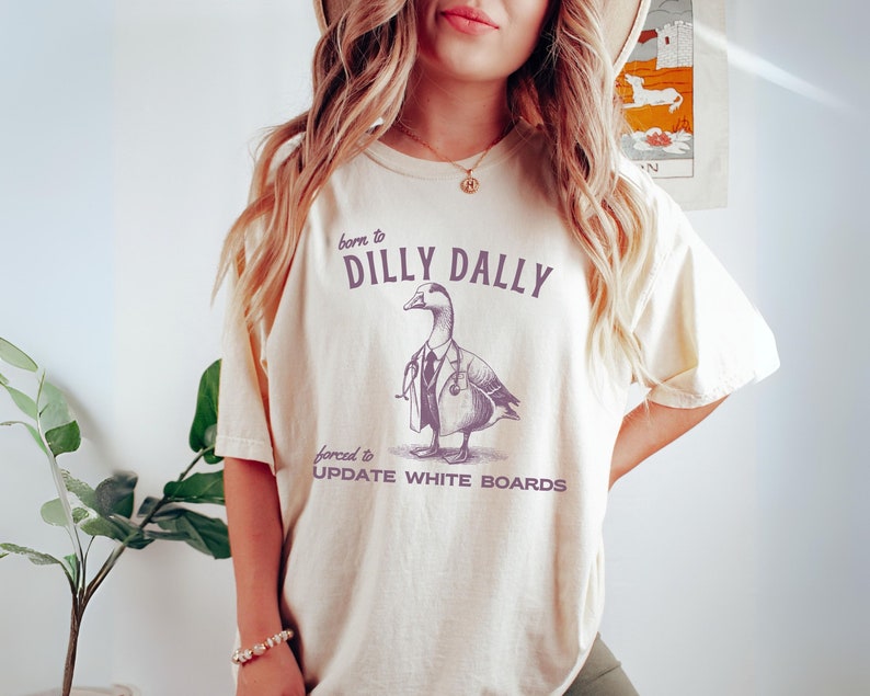 May include: A cream-colored t-shirt with a purple graphic of a doctor duck wearing a stethoscope and the text "born to dilly dally forced to update white boards".