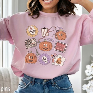 May include: Light pink sweatshirt with a graphic print of various Halloween-themed medical illustrations, including pumpkins, a stethoscope, a skull, and a heart monitor.