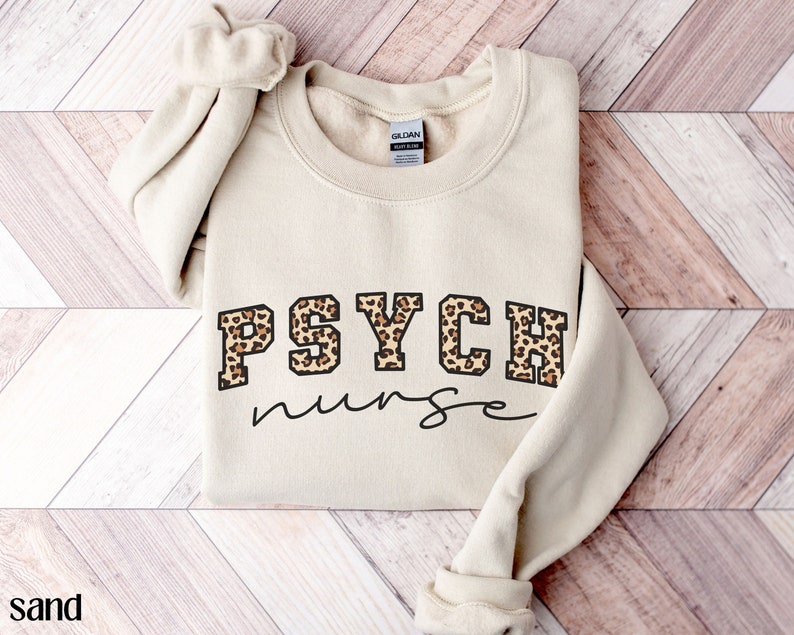 May include: Sand-colored sweatshirt with the words "PSYCH nurse" printed on the front. The word "PSYCH" is in a block letter style with a leopard print pattern. The word "nurse" is in a handwritten font.
