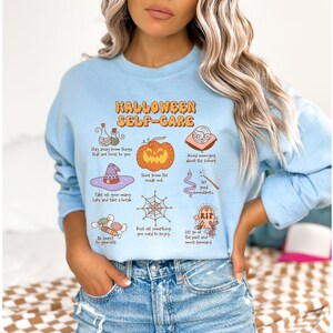May include: Light blue sweatshirt with a Halloween self-care graphic. The graphic features a pumpkin, a witch's hat, a spiderweb, a potion bottle, a tombstone, and a candy cane. The text reads "Halloween Self-Care" and includes phrases like "Stay away from things that are toxic to you", "Shine from the inside out", and "Let go of the past and move forward."