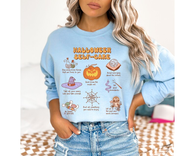 May include: Light blue sweatshirt with a Halloween self-care graphic. The graphic features a pumpkin, a witch's hat, a spiderweb, a potion bottle, a tombstone, and a candy cane. The text reads "Halloween Self-Care" and includes phrases like "Stay away from things that are toxic to you", "Shine from the inside out", and "Let go of the past and move forward."