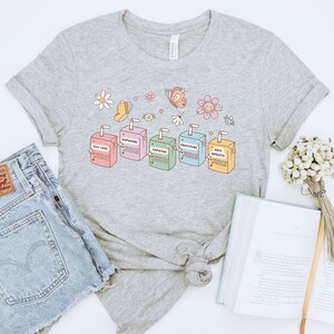 May include: A heather grey t-shirt with a graphic of five colorful juice boxes. The juice boxes are labeled "Self Love Juice", "Dopamine Juice", "Empathy Juice", "Gratitude Juice", and "Anti Anxiety Juice". The graphic also includes butterflies and flowers.