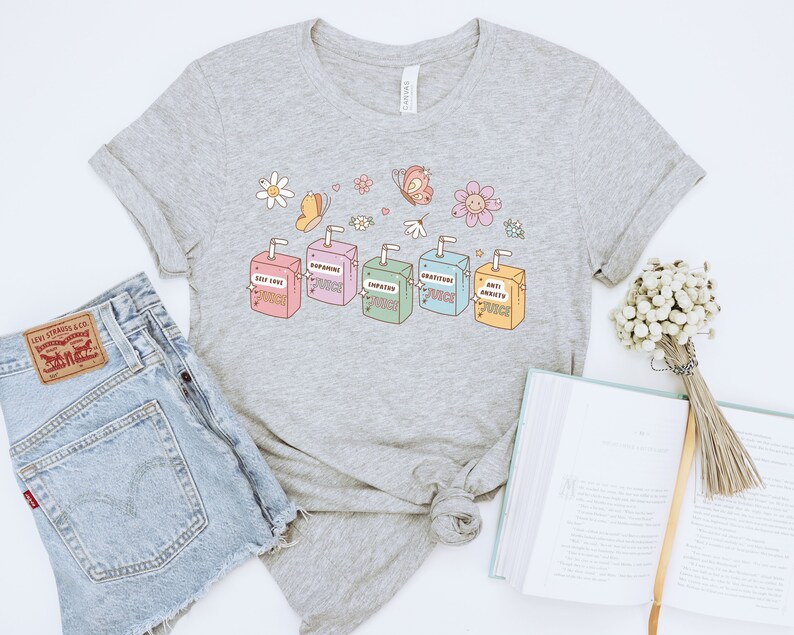 May include: A heather grey t-shirt with a graphic of five colorful juice boxes. The juice boxes are labeled "Self Love Juice", "Dopamine Juice", "Empathy Juice", "Gratitude Juice", and "Anti Anxiety Juice". The graphic also includes butterflies and flowers.