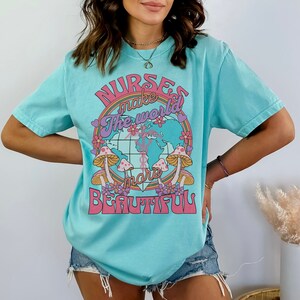 Puede incluir: Una camiseta azul turquesa con un estampado gr&aacute;fico de un globo terr&aacute;queo rodeado de flores y setas. El texto de la camiseta dice "Nurses make the world more beautiful."