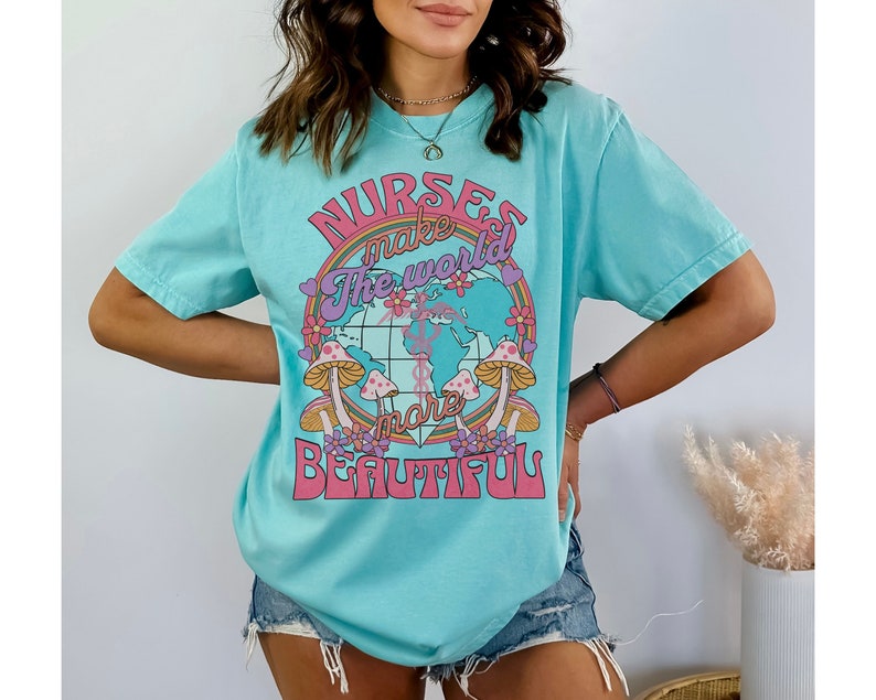 Puede incluir: Una camiseta azul turquesa con un estampado gr&aacute;fico de un globo terr&aacute;queo rodeado de flores y setas. El texto de la camiseta dice "Nurses make the world more beautiful."