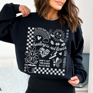 May include: Black sweatshirt with a white graphic print featuring a sun, a flower, a rainbow, and the text "BE YOUR OWN SUNSHINE", "healing is not linear", "FIND JOY IN THE JOURNEY", "DON'T LET THE HARD DAYS WIN", "LOVE YOURSELF", and "988".