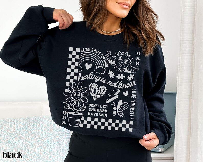 May include: Black sweatshirt with a white graphic print featuring a sun, a flower, a rainbow, and the text "BE YOUR OWN SUNSHINE", "healing is not linear", "FIND JOY IN THE JOURNEY", "DON'T LET THE HARD DAYS WIN", "LOVE YOURSELF", and "988".