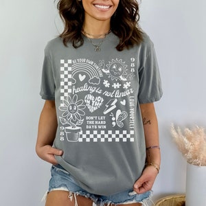 May include: Grey t-shirt with a white graphic design featuring a chequered pattern, a flower, a sun, a butterfly, and text that reads "BE YOUR OWN SUNSHINE", "healing is not linear", "FIND JOY IN THE JOURNEY", "DON'T LET THE HARD DAYS WIN", and "LOVE YOURSELF".