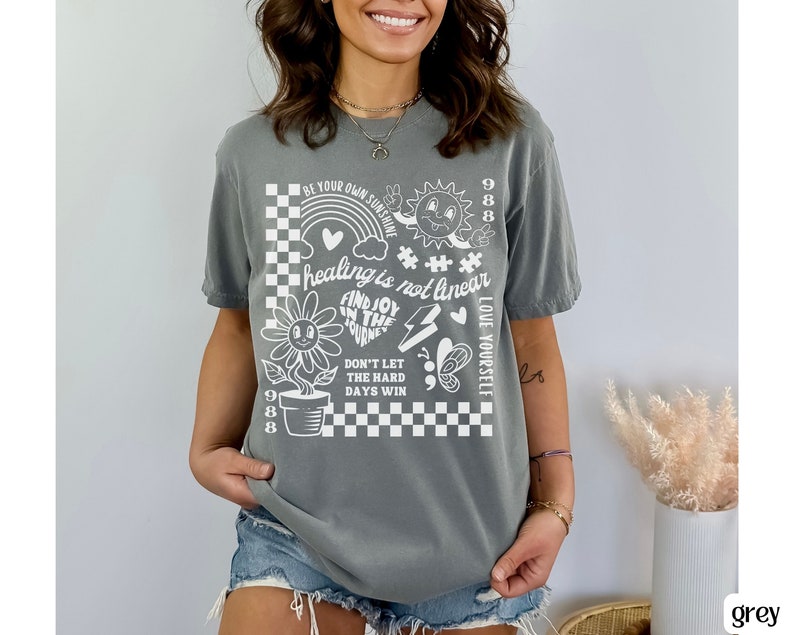May include: Grey t-shirt with a white graphic design featuring a chequered pattern, a flower, a sun, a butterfly, and text that reads "BE YOUR OWN SUNSHINE", "healing is not linear", "FIND JOY IN THE JOURNEY", "DON'T LET THE HARD DAYS WIN", and "LOVE YOURSELF".