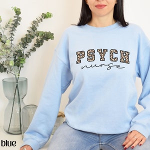May include: Light blue sweatshirt with the text "PSYCH nurse" in a combination of leopard print and black script. The sweatshirt is a crew neck style and is made of a soft material.