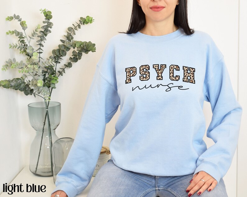 May include: Light blue sweatshirt with the text "PSYCH nurse" in a combination of leopard print and black script. The sweatshirt is a crew neck style and is made of a soft material.