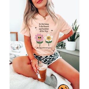 May include: A pink t-shirt with a graphic of two cartoon flowers with faces. The text on the shirt reads "It's Not Going To Be Flowers and Sunshine All the Time. But they will be back again."