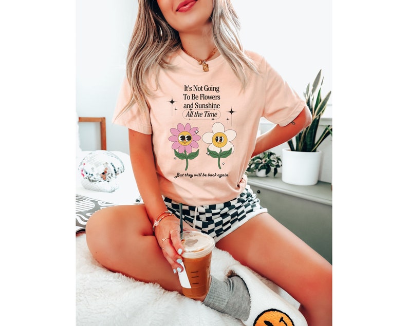 May include: A pink t-shirt with a graphic of two cartoon flowers with faces. The text on the shirt reads "It's Not Going To Be Flowers and Sunshine All the Time. But they will be back again."