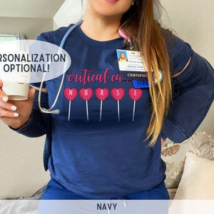 Pu&ograve; includere: Maglietta blu navy con il testo "critical care nurse" e grafiche di lecca-lecca rosa. Il testo "CERTIFIED RN" &egrave; su un cartellino identificativo.
