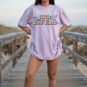 May include: A light purple t-shirt with the text "SSR's and Good Vibes" in a retro font. The graphic includes a rainbow, sun, and other elements. The shirt is a relaxed fit.